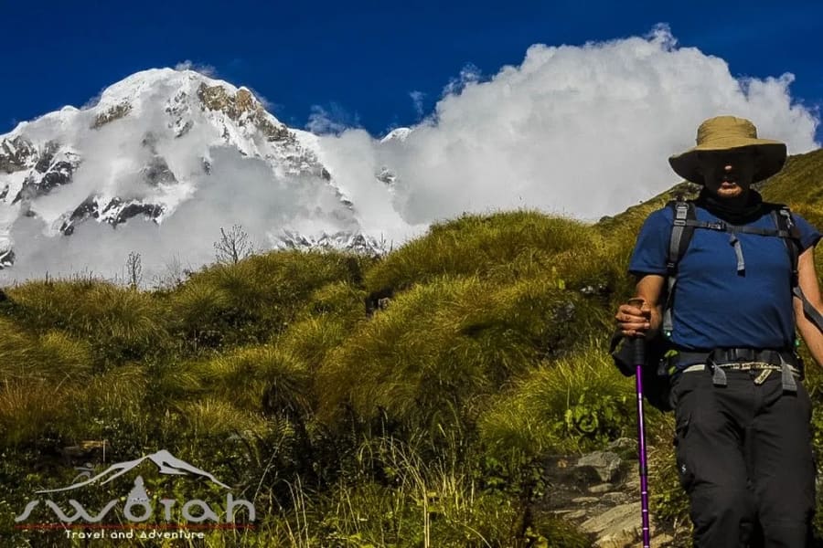 A trekker in a sun hat on a grassy Annapurna trail below the cloud-wrapped Annapurna South peak
